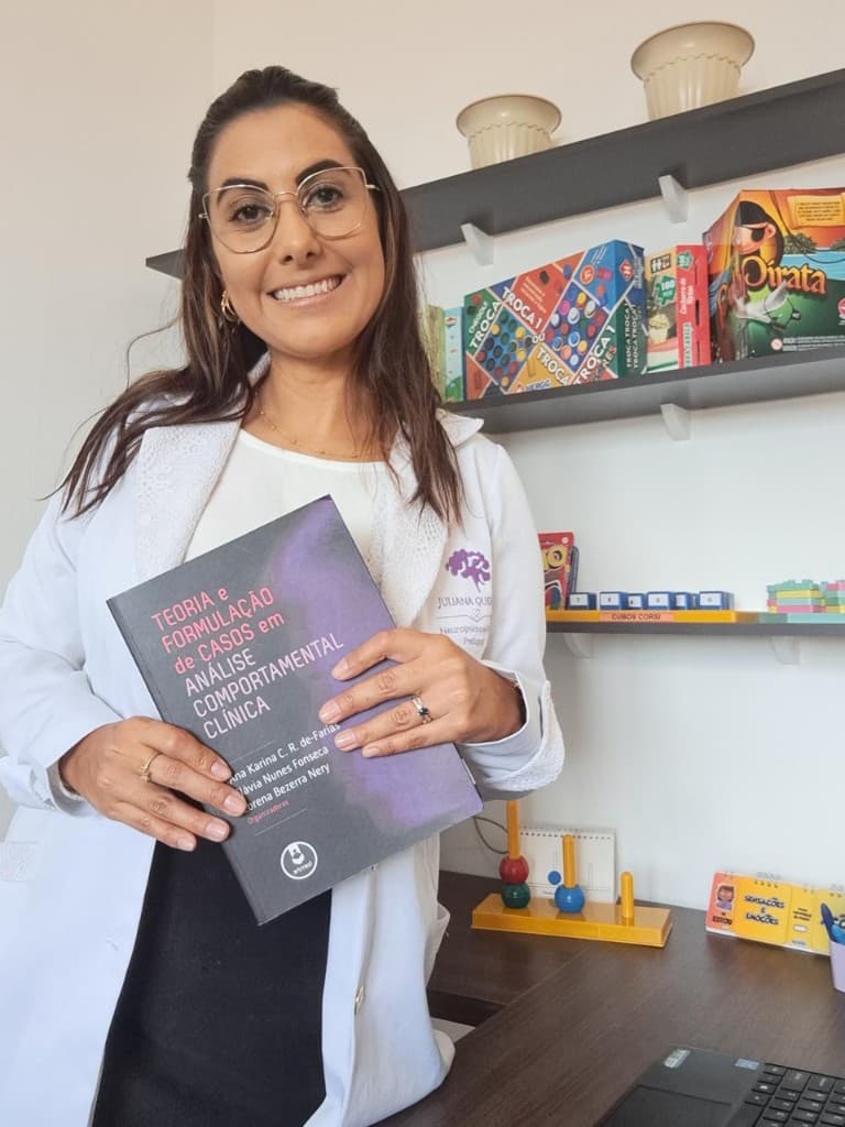 Juliana Queiroz no consultório, de jaleco branco, segurando um livro de formulação de casos em análise do comportamento clínica.