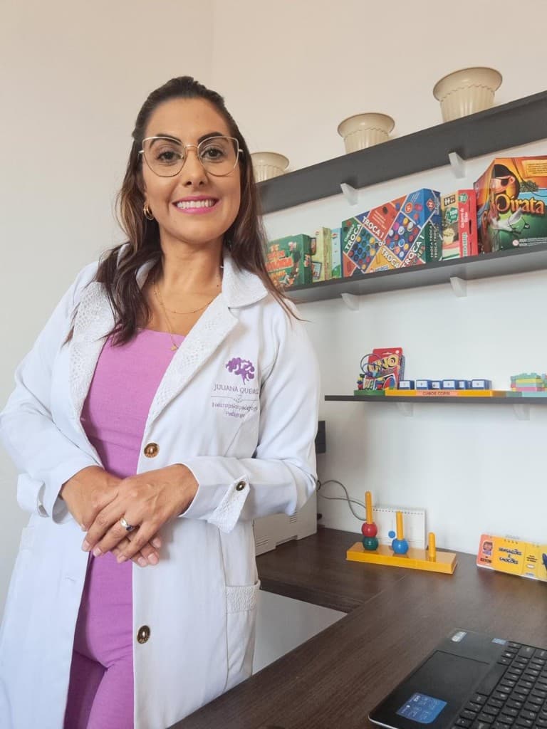 Juliana Queiroz, neuropsicopedagoga, sorrindo no consultório. Ela usa jaleco branco com logo roxo e óculos dourados; ao fundo, prateleiras com jogos e materiais pedagógicos.
