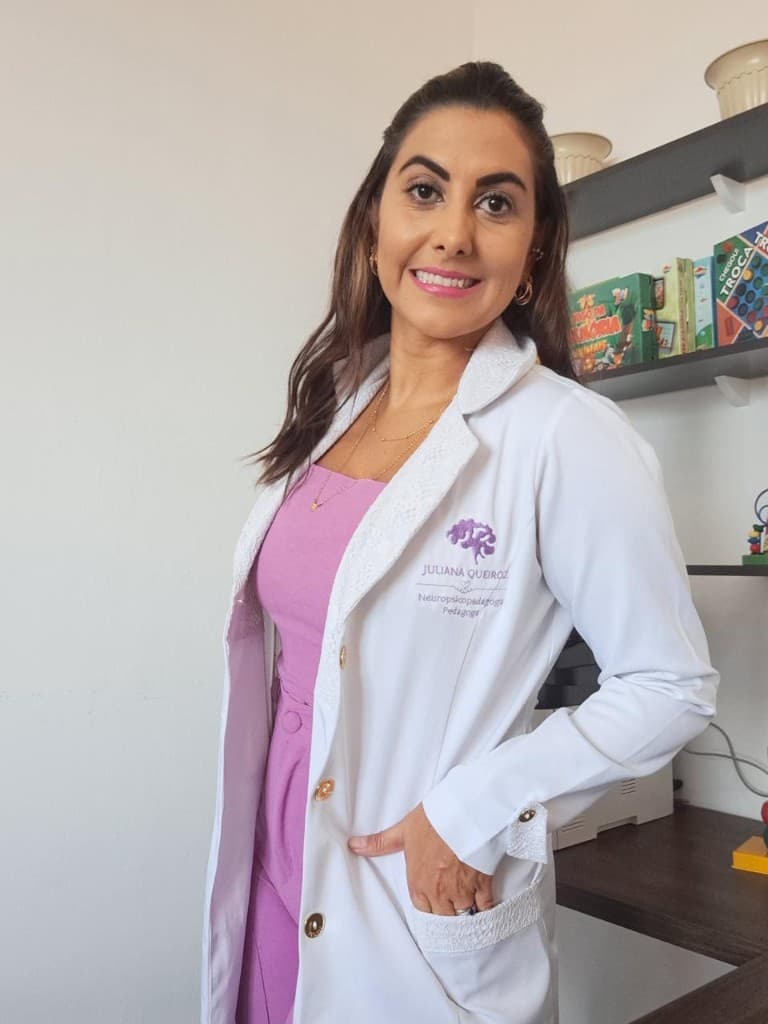Juliana Queiroz de jaleco branco com logo roxo, em ambiente de consultório com materiais pedagógicos ao fundo.